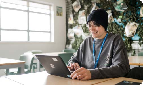 Student sat at a desk in a classroom wearing a beanie and college lanyard, looking down and smiling whilst working on a laptop